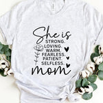 White graphic T-shirt reading ‘She is strong, loving, warm, fearless, patient, selfless—mom’ styled flat-lay with tan fedora, denim shorts, and greenery.