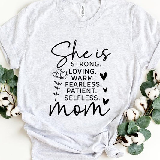 White graphic T-shirt reading ‘She is strong, loving, warm, fearless, patient, selfless—mom’ styled flat-lay with tan fedora, denim shorts, and greenery.