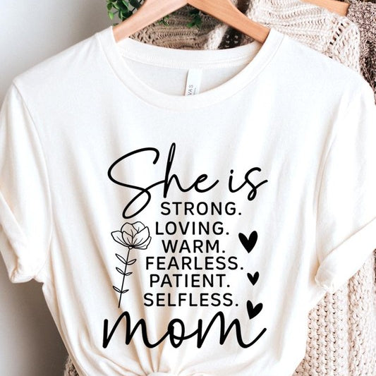 White graphic T-shirt reading ‘She is strong, loving, warm, fearless, patient, selfless—mom’ styled flat-lay with tan fedora, denim shorts, and greenery.