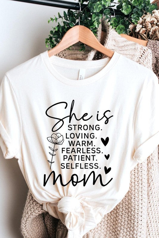 White graphic T-shirt reading ‘She is strong, loving, warm, fearless, patient, selfless—mom’ styled flat-lay with tan fedora, denim shorts, and greenery.