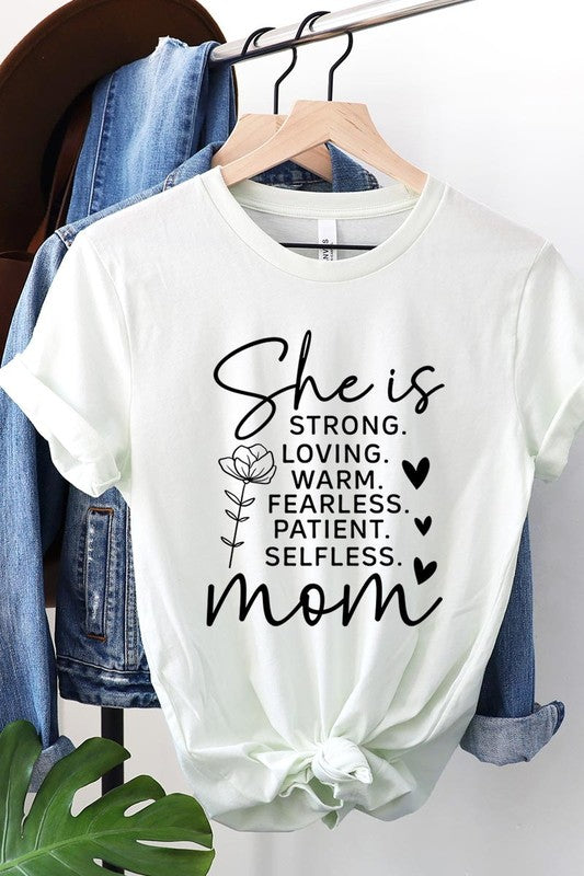 White graphic T-shirt reading ‘She is strong, loving, warm, fearless, patient, selfless—mom’ styled flat-lay with tan fedora, denim shorts, and greenery.