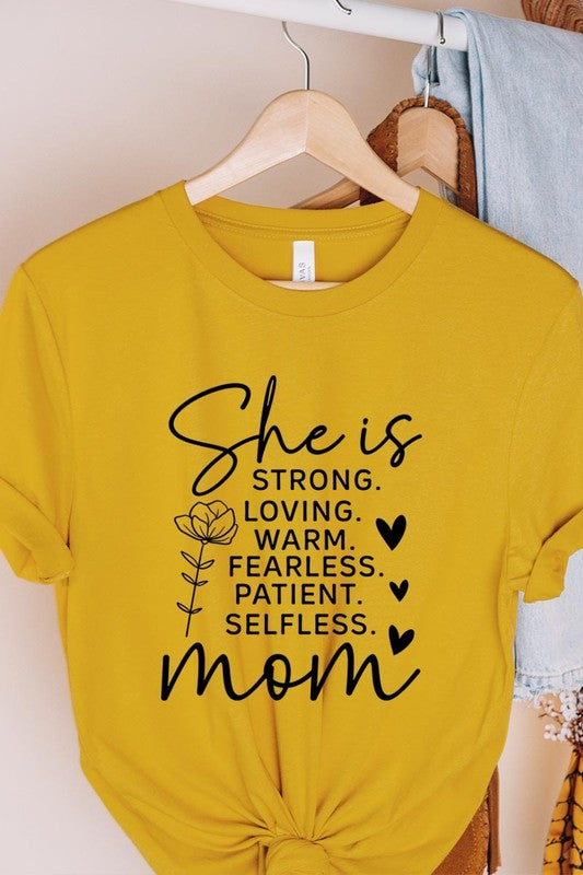 Yellow graphic T-shirt reading ‘She is strong, loving, warm, fearless, patient, selfless—mom’ styled flat-lay with tan fedora, denim shorts, and greenery.