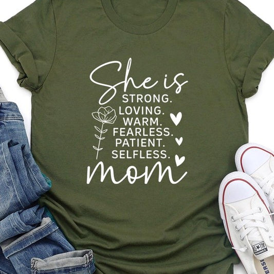 Army green graphic T-shirt reading ‘She is strong, loving, warm, fearless, patient, selfless—mom’ styled flat-lay with tan fedora, denim shorts, and greenery.