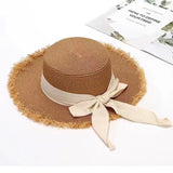 Cream straw sun hat with frayed brim and long grosgrain bow ribbon tied at the back, displayed against a soft floral backdrop.