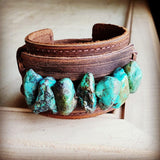 Handcrafted brown leather cuff bracelet featuring raw turquoise stones, displayed on a rustic wooden surface.