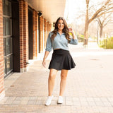 Woman styling a black high-waisted tummy-control skort with built-in shorts and flared A-line overlay, showcasing slimming athletic fit for tennis, golf or everyday wear.