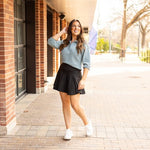 Woman styling a black high-waisted tummy-control skort with built-in shorts and flared A-line overlay, showcasing slimming athletic fit for tennis, golf or everyday wear.