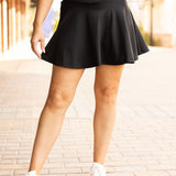 Woman styling a black high-waisted tummy-control skort with built-in shorts and flared A-line overlay, showcasing slimming athletic fit for tennis, golf or everyday wear.