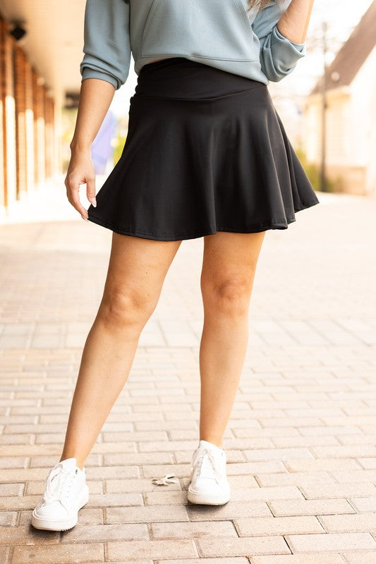 Woman styling a black high-waisted tummy-control skort with built-in shorts and flared A-line overlay, showcasing slimming athletic fit for tennis, golf or everyday wear.