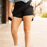 Woman styling a black high-waisted tummy-control skort with built-in shorts and flared A-line overlay, showcasing slimming athletic fit for tennis, golf or everyday wear.