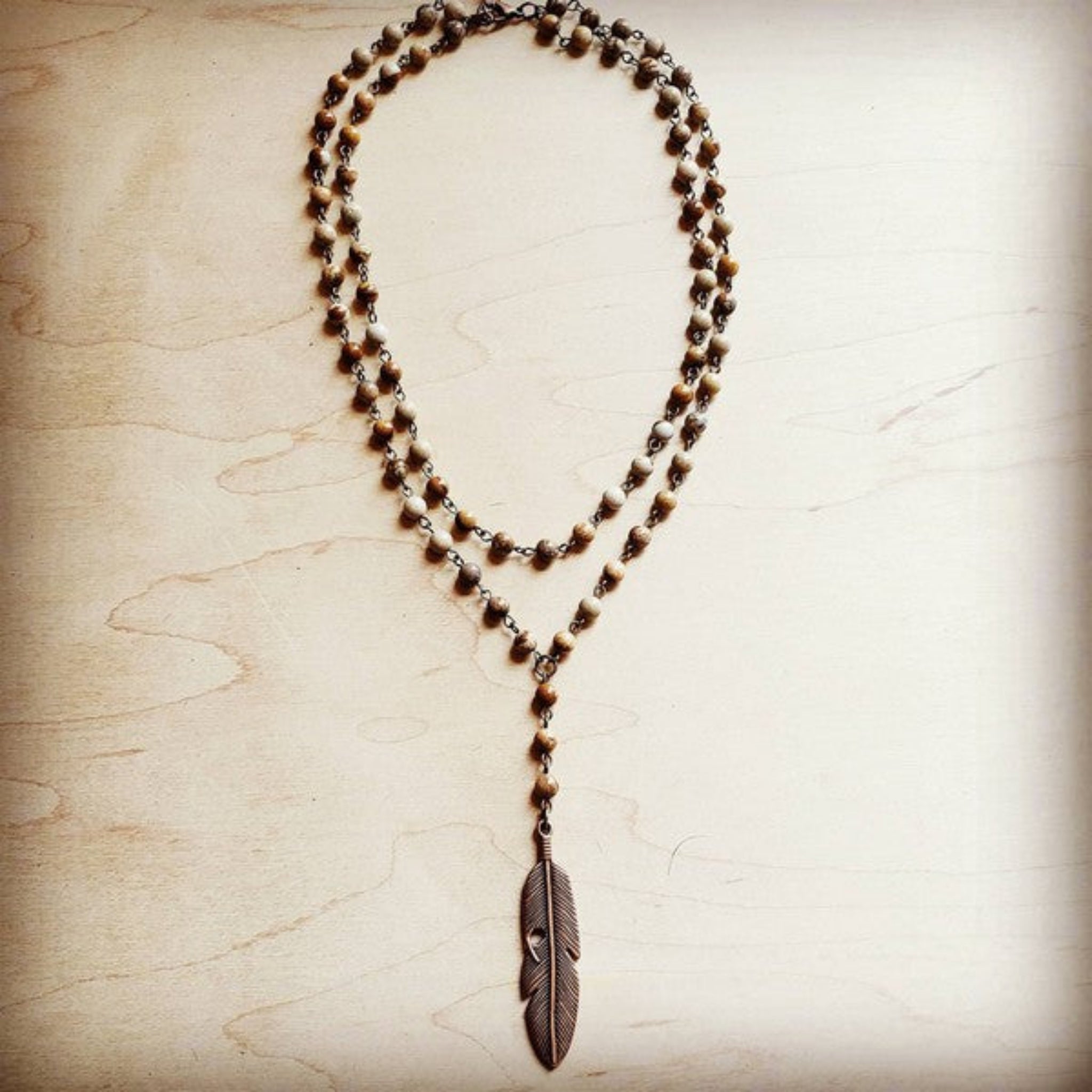 Layered beaded necklace with brown and cream stones and bronze feather pendant, displayed on light wooden background.