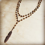 Layered beaded necklace with brown and cream stones and bronze feather pendant, displayed on light wooden background.