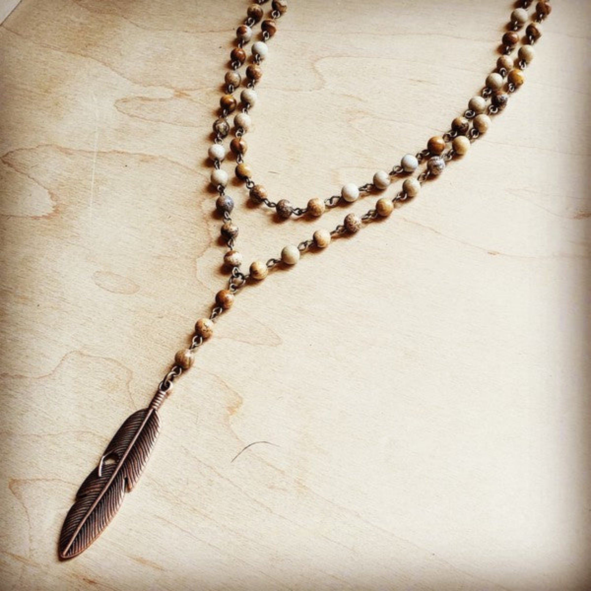 Layered beaded necklace with brown and cream stones and bronze feather pendant, displayed on light wooden background.
