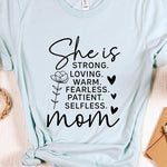 Graphic T-shirt reading ‘She is strong, loving, warm, fearless, patient, selfless—mom’ styled flat-lay with tan fedora, denim shorts, and greenery.