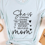 Graphic T-shirt reading ‘She is strong, loving, warm, fearless, patient, selfless—mom’ styled flat-lay with tan fedora, denim shorts, and greenery.