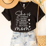 Black graphic T-shirt reading ‘She is strong, loving, warm, fearless, patient, selfless—mom’ styled flat-lay with tan fedora, denim shorts, and greenery.