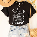 Black graphic T-shirt reading ‘She is strong, loving, warm, fearless, patient, selfless—mom’ styled flat-lay with tan fedora, denim shorts, and greenery.
