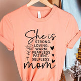 Creamsicle Orange graphic T-shirt reading ‘She is strong, loving, warm, fearless, patient, selfless—mom’ styled flat-lay with tan fedora, denim shorts, and greenery.