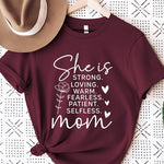 Crimson Red graphic T-shirt reading ‘She is strong, loving, warm, fearless, patient, selfless—mom’ styled flat-lay with tan fedora, denim shorts, and greenery.