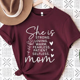 Crimson Red graphic T-shirt reading ‘She is strong, loving, warm, fearless, patient, selfless—mom’ styled flat-lay with tan fedora, denim shorts, and greenery.