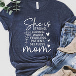Graphic T-shirt reading ‘She is strong, loving, warm, fearless, patient, selfless—mom’ styled flat-lay with tan fedora, denim shorts, and greenery.