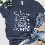 Graphic T-shirt reading ‘She is strong, loving, warm, fearless, patient, selfless—mom’ styled flat-lay with tan fedora, denim shorts, and greenery.