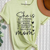 Light Green graphic T-shirt reading ‘She is strong, loving, warm, fearless, patient, selfless—mom’ styled flat-lay with tan fedora, denim shorts, and greenery.