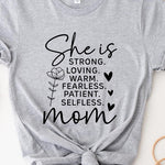 Light Grey graphic T-shirt reading ‘She is strong, loving, warm, fearless, patient, selfless—mom’ styled flat-lay with tan fedora, denim shorts, and greenery.