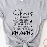 Light Grey graphic T-shirt reading ‘She is strong, loving, warm, fearless, patient, selfless—mom’ styled flat-lay with tan fedora, denim shorts, and greenery.