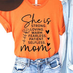 Orange graphic T-shirt reading ‘She is strong, loving, warm, fearless, patient, selfless—mom’ styled flat-lay with tan fedora, denim shorts, and greenery.