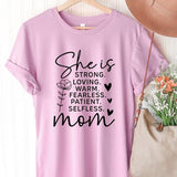 Pink graphic T-shirt reading ‘She is strong, loving, warm, fearless, patient, selfless—mom’ styled flat-lay with tan fedora, denim shorts, and greenery.