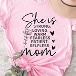 Pink graphic T-shirt reading ‘She is strong, loving, warm, fearless, patient, selfless—mom’ styled flat-lay with tan fedora, denim shorts, and greenery.