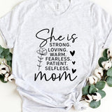 White graphic T-shirt reading ‘She is strong, loving, warm, fearless, patient, selfless—mom’ styled flat-lay with tan fedora, denim shorts, and greenery.