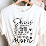 White graphic T-shirt reading ‘She is strong, loving, warm, fearless, patient, selfless—mom’ styled flat-lay with tan fedora, denim shorts, and greenery.