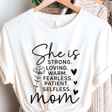 White graphic T-shirt reading ‘She is strong, loving, warm, fearless, patient, selfless—mom’ styled flat-lay with tan fedora, denim shorts, and greenery.