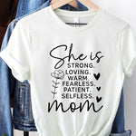 White graphic T-shirt reading ‘She is strong, loving, warm, fearless, patient, selfless—mom’ styled flat-lay with tan fedora, denim shorts, and greenery.