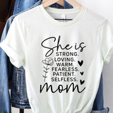 White graphic T-shirt reading ‘She is strong, loving, warm, fearless, patient, selfless—mom’ styled flat-lay with tan fedora, denim shorts, and greenery.