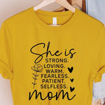 Yellow graphic T-shirt reading ‘She is strong, loving, warm, fearless, patient, selfless—mom’ styled flat-lay with tan fedora, denim shorts, and greenery.