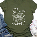Army green graphic T-shirt reading ‘She is strong, loving, warm, fearless, patient, selfless—mom’ styled flat-lay with tan fedora, denim shorts, and greenery.