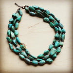 Chunky three-strand turquoise statement necklace with natural raw-cut stones, displayed on a light wooden background.