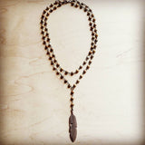 Layered beaded necklace with brown and cream stones and bronze feather pendant, displayed on light wooden background.