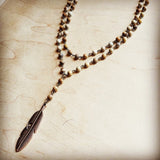 Layered beaded necklace with brown and cream stones and bronze feather pendant, displayed on light wooden background.